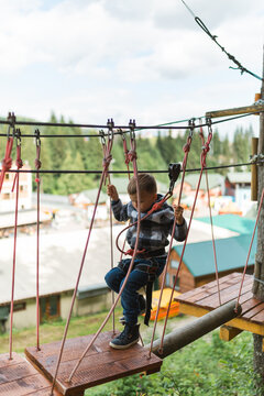 Focused child boy navigating through tricky rope bridge obstacle at high ropes course in woods.