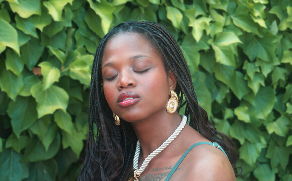 Serene young black woman with braided hair and gold earrings closing eyes in front of green ivy wall.
