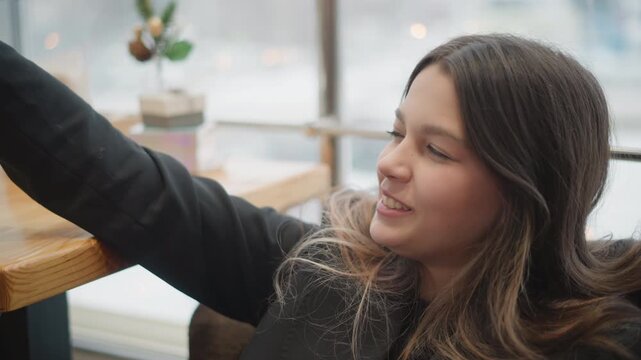 Young woman seated beside bright window in cozy cafe extends arm across wooden table while gazing aside with focused expression, warm casual style, soft natural light, and relaxed urban mood