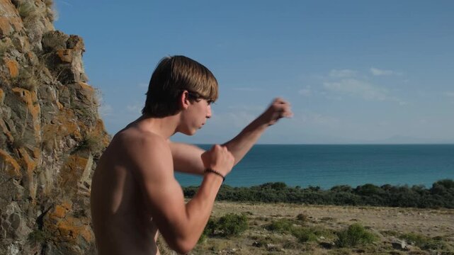 Shirtless young athlete with a muscular defined body practicing shadow boxing on the seashore among rocks, throwing punches forward, solo outdoor martial arts training and combat sports