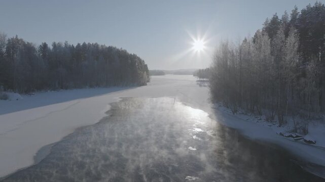 Aerial drone footage flying over frozen river with sun glare in winter forest landscape