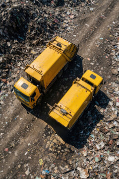 Two yellow garbage trucks operate at a vast landfill site. Trucks collect and haul refuse across dirt roads among large piles of waste. Aerial view shows industrial waste management process.