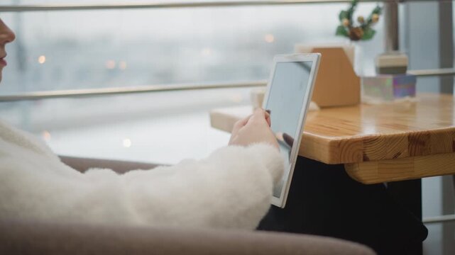 Close over shoulder view shows person seated beside window at wooden caf table, tapping tablet screen with index finger while holding device with other hand in soft daylight, with tablet, hand, tap