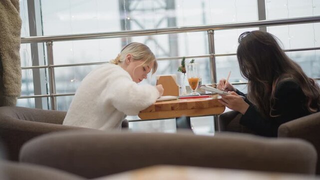 Two women sit across from each other at wooden caf table, both leaning forward and concentrating on objects on table; light-haired woman reaches toward paper or device while dark-haired woman looks