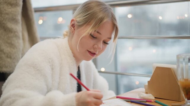 Close three-quarter view of young girl leaning over table and drawing with colored pencil, visible hand movement and joyful focused expression showing engaged creativity in cozy indoor daylight