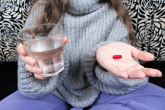 Child in warm knitted sweater holding a red capsule pill in open palm and a glass of water, ready to take medicine at home
