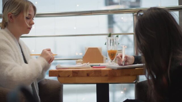 Blond youth in white fuzzy sweater sits across companion with long dark hair at round wooden table inside bright cafe, reaching toward plate, handling food, and sharing relaxed conversation in soft