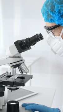 Senior researcher in protective gear calibrates microscope while examining sample, researching breakthrough medical research in sterile laboratory conditions. Medicine and science concept