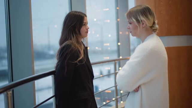 Two young adults stand facing each other beside glass railing inside modern office, with city skyline visible through window, with conversation, speaking, listening, interaction detail