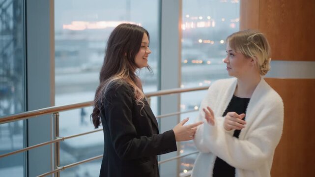 Two young women stand beside floor to ceiling windows and metal railing in bright modern interior, sharing warm casual conversation as one gestures lightly toward other in relaxed workplace setting