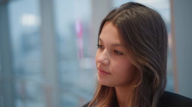Young woman turns head left while speaking softly, mouth slightly open and eyes looking off camera, showing shift from quiet contemplation toward gentle engagement in bright indoor daylight near glass