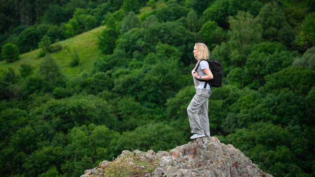 Blonde woman with a backpack standing on a rocky peak. Female hiker looking at a green mountain forest. Outdoor adventure, hike and travel concept