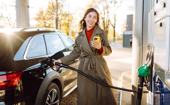 Smiling woman fills up car at gas station on a sunny day. Stylish woman stands near car at gas station, holding a phone in her hand. Concept: gas stations, transportation, and service.