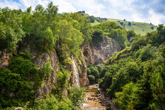 Waterfalls down a rocky cliff in a green mountain gorge near Kislovodsk, Russia. Tourists walking on a path in a summer landscape, nature concept