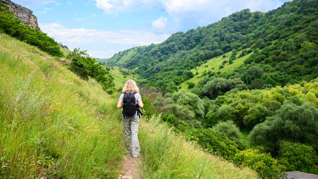 Blonde woman hiking on a mountain trail with a backpack. Female hiker walking along a grassy slope overlooking a lush green valley. Outdoor adventure and active lifestyle concept