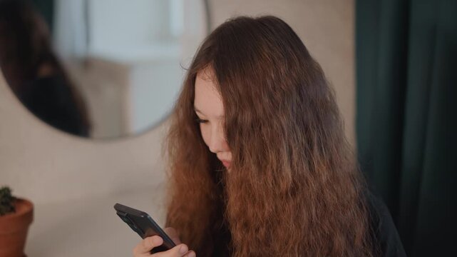Medium close view shows young woman in bedroom dressing area, head tilted down, hands steady on smartphone, eyes fixed on screen, suggesting scrolling or texting with quiet, concentrated attention