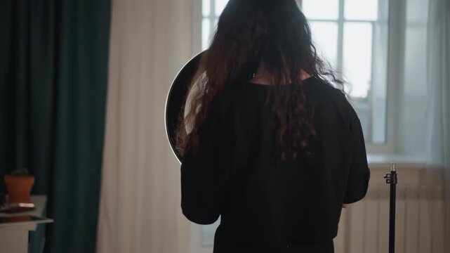Rear view of woman standing beside circular LED light near tall window, holding smartphone at chest level while checking framing and light balance in calm home studio with neutral decor and soft