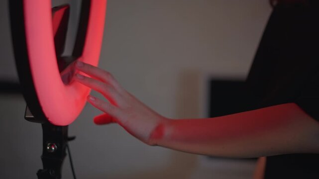 Close-up hand and forearm adjusting circular ring light rim while colored glow shifts through blue, pink, red, neutral white, creating tactile studio lighting change in dark setting