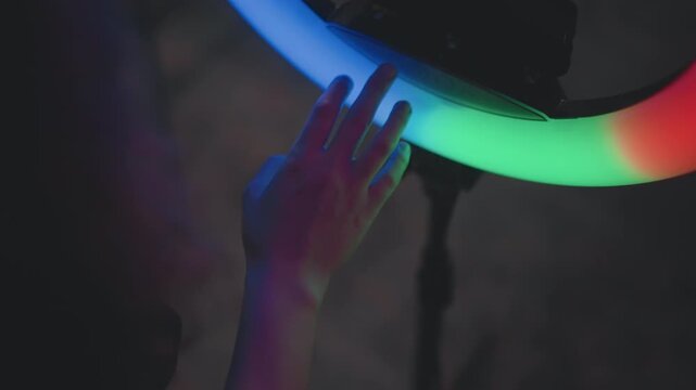 Close up of hand touching partial circular ring light as colors shift through orange, green, blue, purple, and white segments, with soft glow, gentle contact, and moody studio atmosphere