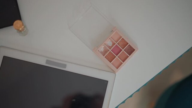 Overhead view shows arm reaching across bright white desk toward tilted tablet beside open eyeshadow palette with visible color pans, under soft daylight and gentle shadows in clean minimal workspace