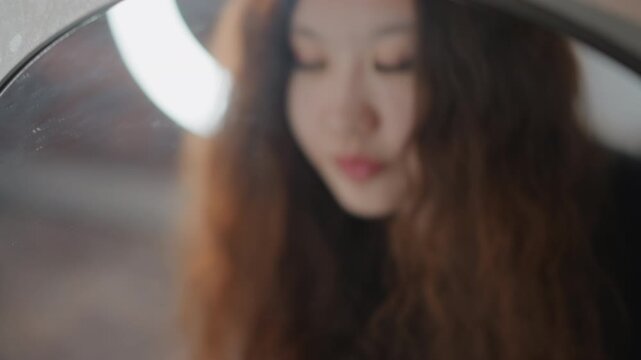 Close-up motion shows young woman carefully adjusting false eyelashes while looking into mirror, gentle fingertip movement near eye and slight head tilt revealing focused beauty routine