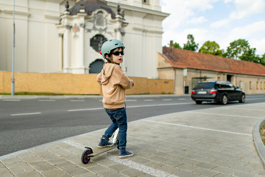 Young boy wearing a safety helmet rides a kick scooter on a city sidewalk. Active childhood, urban mobility and road safety for children.