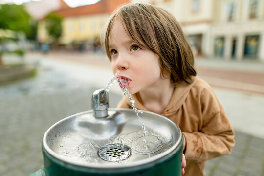 Playful young boy having fun with a public drinking fountain in the city. Laughing child splashes with fresh water outdoors during a playful urban summer moment.