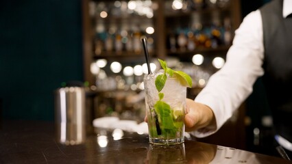 Bartender hand offers mojito in tall glass with mint leaves lime wedge. Dark bar counter, bokeh lights, cool tones. Perfect for nightlife, beverage, hospitality concepts © Ruslan