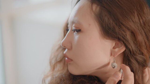 Close view of young woman in profile gently turning head while lifting hand to adjust dangling earring and brush loose wavy hair; soft warm natural light from left creates calm intimate beauty mood