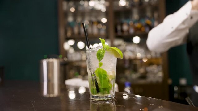 Bartender hand offers mojito in tall glass with mint leaves lime wedge. Dark bar counter, bokeh lights, cool tones. Perfect for nightlife, beverage, hospitality concepts