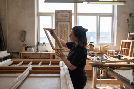 Female carpenter holds part of antique window frame in hands examining quality of wooden part, professional woman woodworker checking condition of wood and planning stages of restoration work.