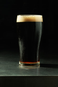 Full pint of dark craft beer served in a chilled glass with a creamy foam head against a black background. Premium stout or porter beer with visible condensation, dramatic studio lighting