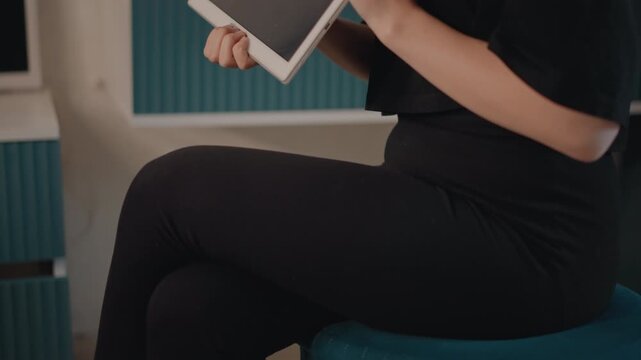 Profile view of woman from chest up holding dark screen tablet in one hand and pointing with index finger, slight forward lean and focused expression suggest interaction, explanation, or guidance