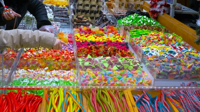 Colorful candy stall selling gummy sweets at market. Large assortment of colorful jelly sweets and candies on display at a street market stall, with people choosing different types of sugary treats
