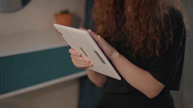 Medium closeup threequarter view of young woman holding tablet with both hands, actively tapping and scrolling while looking downward with focused calm expression in modern indoor setting