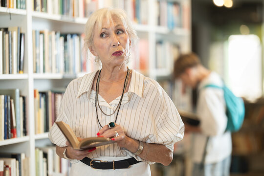 Interested elderly female visitor holding a book in public library with large stock
