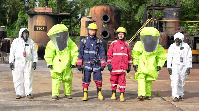 Hazard material protective suit chemical suit gas mask firefighter team at industrial site emergency response training drill, worker group walks past tanks on concrete yard, focused risk control