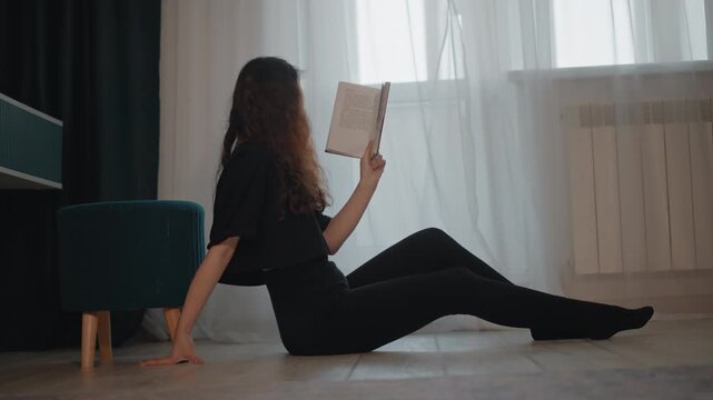 Young woman sits on floor leaning back on one hand while holding book upright at eye level, indicating active reading with slight torso rotation toward pages
