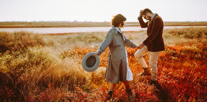 Young man and woman walk together across bright grassy field at sunset. A loving couple in stylish clothing enjoys nature and time together outdoors. Concept of love and relaxation.