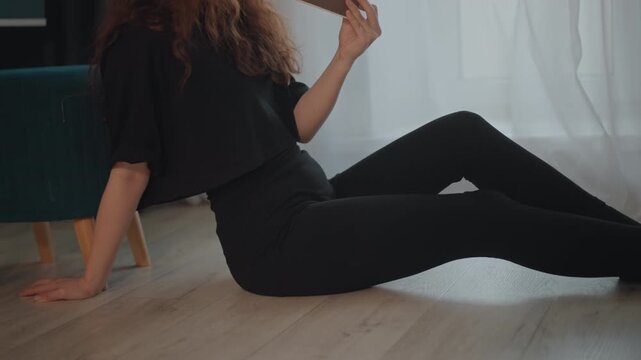 Woman sits on light wood floor beside sheer curtain-covered window, legs extended and one knee bent, holding rectangular object near face as she examines it in soft daylight with relaxed posture