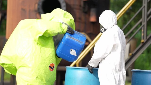 Hazardous waste chemical spill cleanup with protective suit respirator industrial barrel response, workers transfer liquid from jerrycan into drum for contamination control and safety inspection
