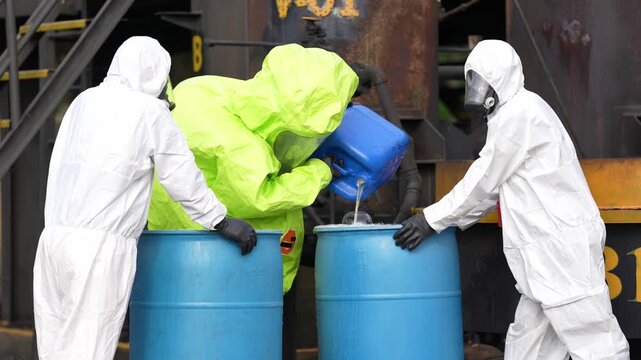 Hazardous material suit chemical spill hazardous waste industrial site safety protective gear workers pour liquid into drum during decontamination cleanup emergency response