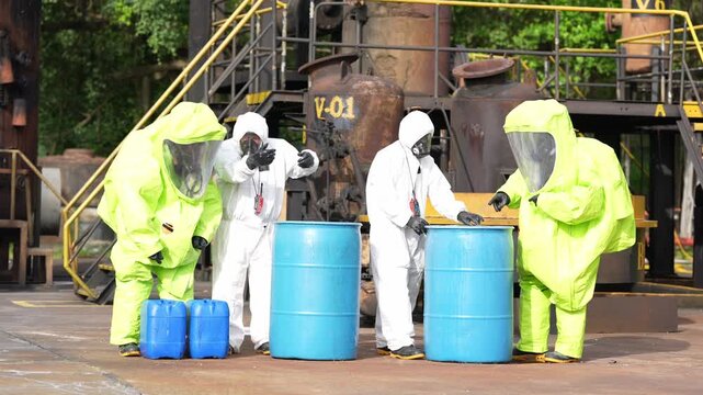 Hazmat chemical spill response worker in protective suit inspects blue barrel at factory industrial site, emergency cleanup team handles hazardous waste drums, safety inspection and decontamination