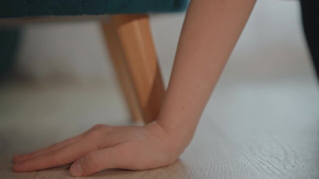 Low-angle view shows hand and forearm sliding across warm hardwood floor beneath furniture leg, fingers and palm touching textured surface while wrist bends during careful reaching motion in quiet