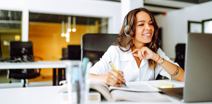 A woman wearing a headset works at a desk with a laptop. A young woman in a modern office works with a notepad and laptop on a sunny day. Support concept, office, employee.