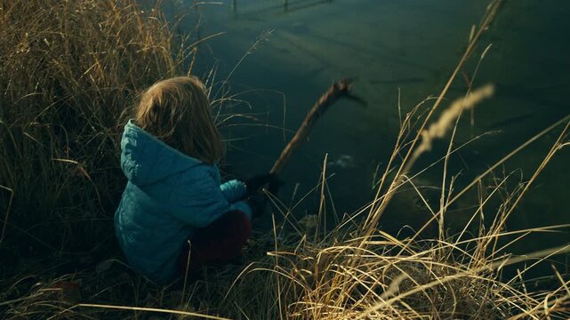 High angle view of boy crouching in dried grass and poking a stick into a frozen river