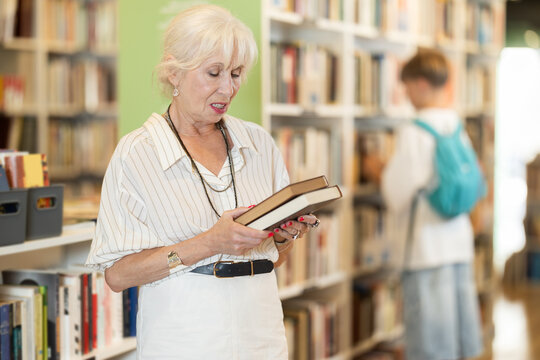 Elderly woman stands in bookstore, carefully browsing shelves and selecting book of interest
