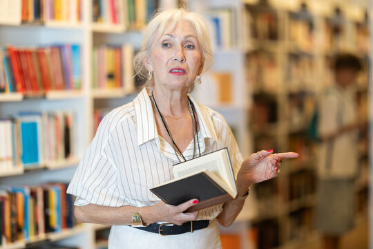 Focused mature lady browsing through literature volumes on shelves in cozy bookshop