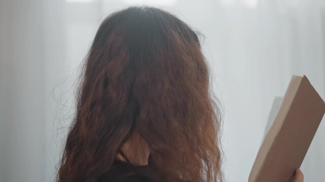 Person shown from behind with long curly brown hair holding rectangular board or canvas in right hand while slightly turning head toward object, suggesting inspection, preparation, and calm creative