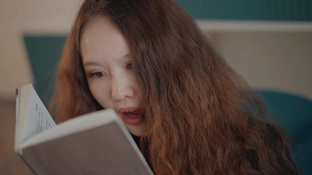 Close view of young woman reading book indoors with steady downward gaze, small head tilt and gently pursed lips, showing concentration, calm reflection and quiet learning mood, with woman, reading